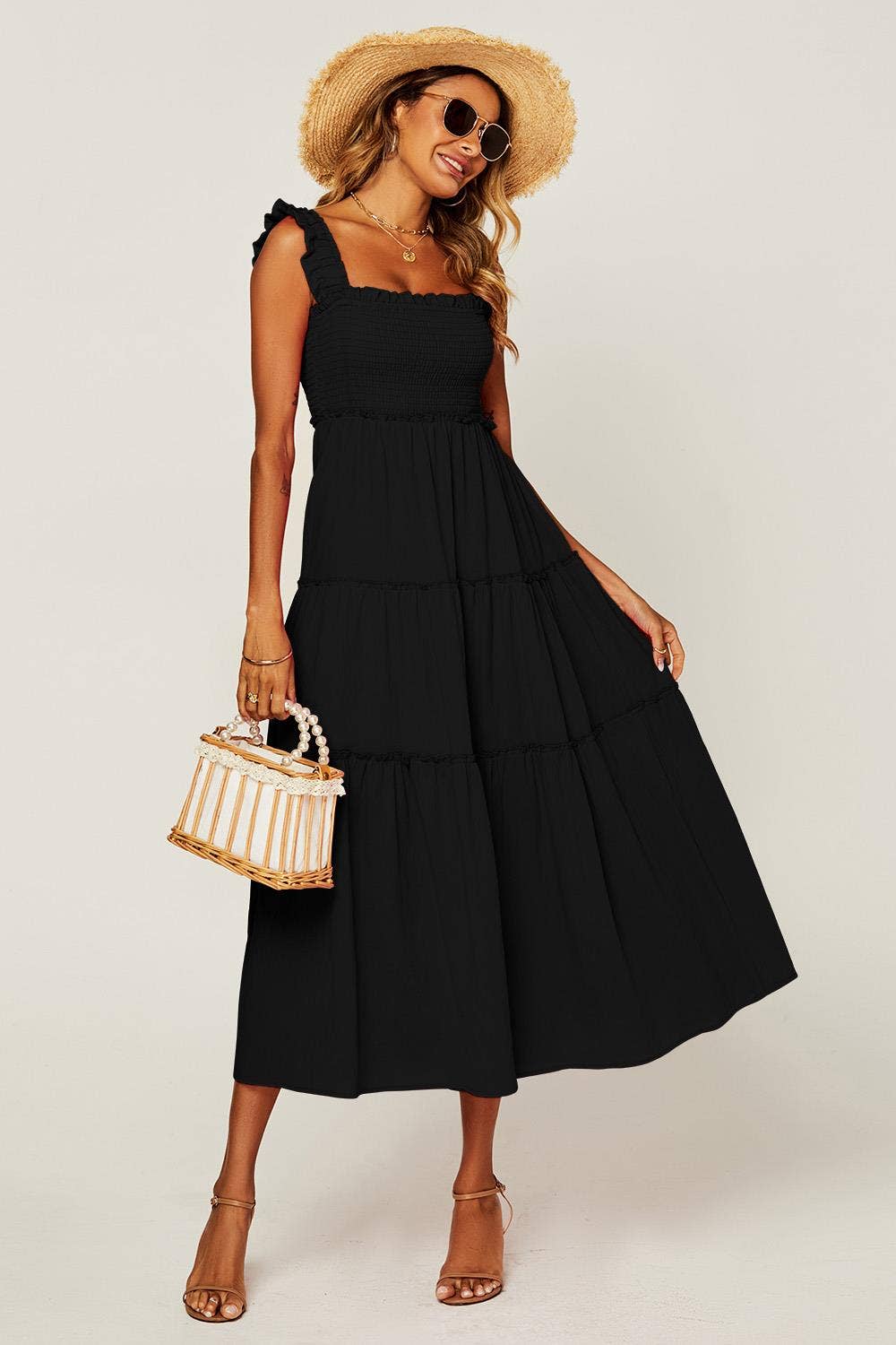A woman modeling a black tiered midi dress with ruffled shoulder straps, styled with a straw hat, sunglasses, a woven handbag, and strappy heels against a light gray studio background.