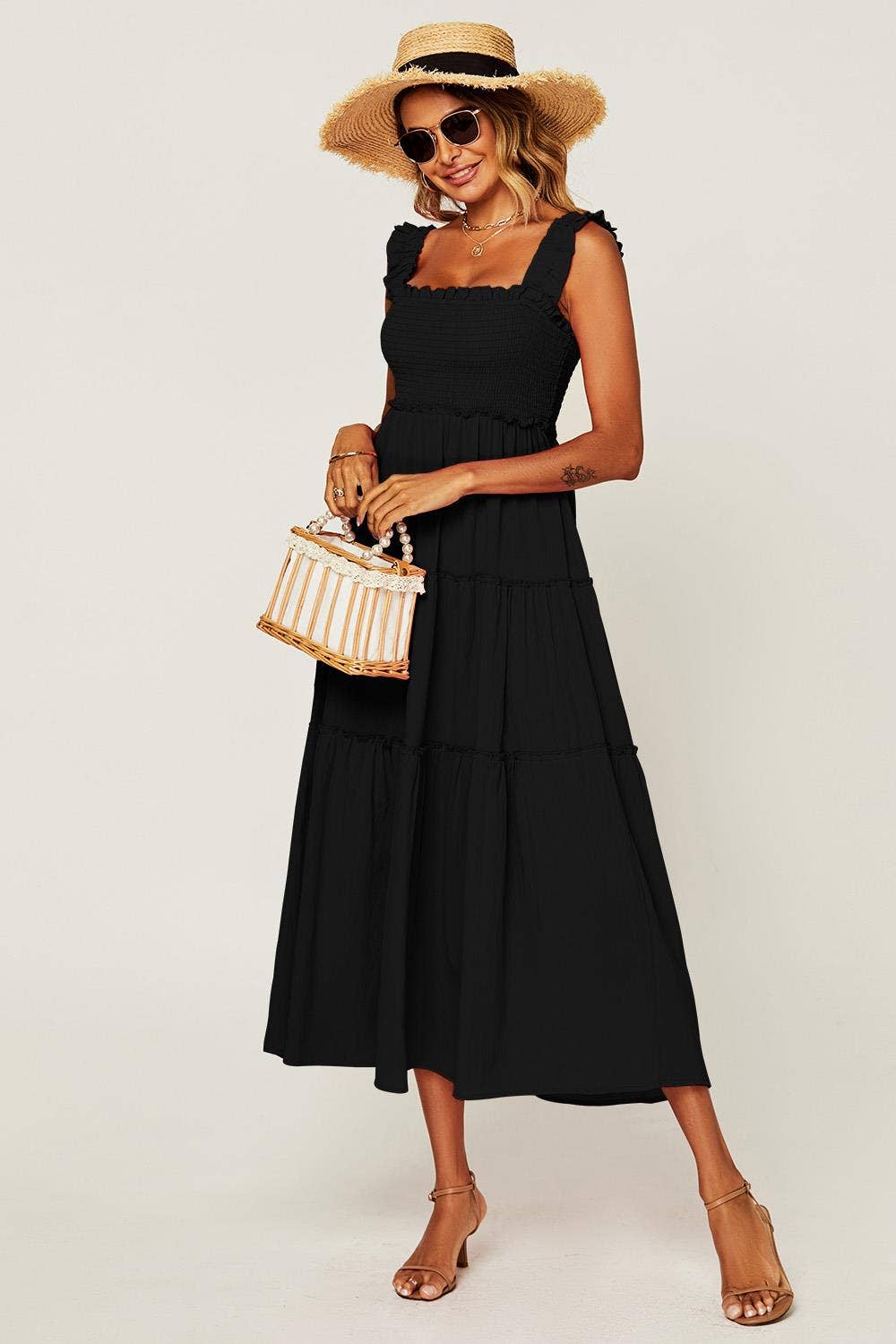 A woman modeling a sleeveless black midi dress with a tiered skirt, styled with a straw hat, sunglasses, a striped handbag, and tan heeled sandals against a plain light background.