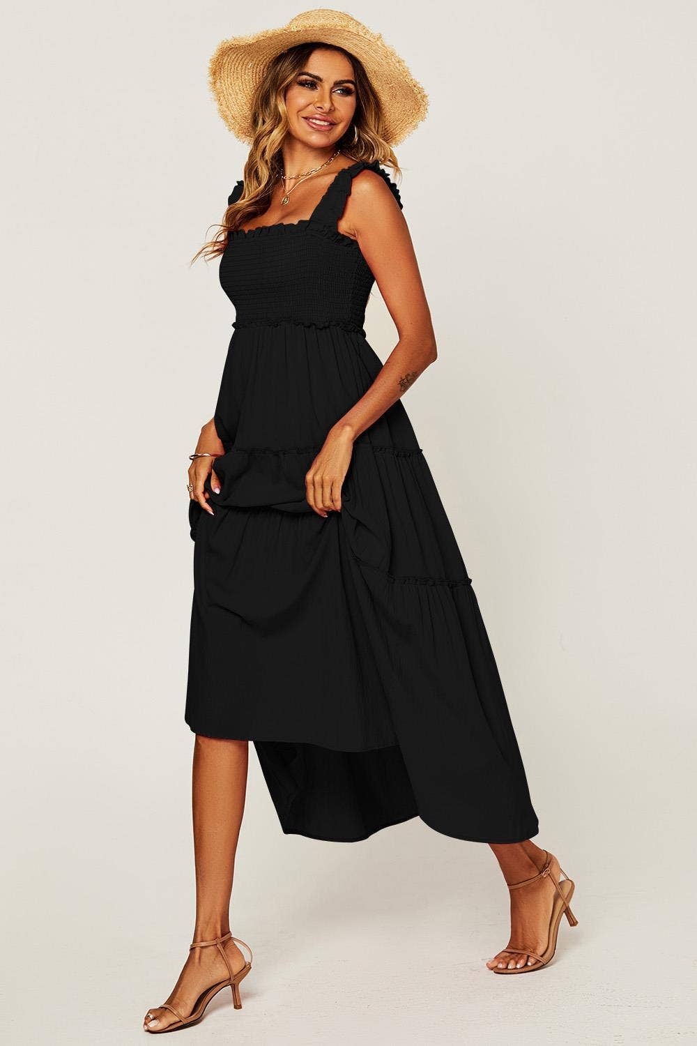 A woman modeling a black midi dress with a tiered skirt and ruffled shoulder straps, styled with a straw hat and tan heeled sandals against a light gray studio background.
