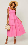 A woman models a bright pink tiered midi dress with ruffled shoulder straps, styled with a straw hat, sunglasses, a woven handbag, and tan heeled sandals against a light gray studio background.