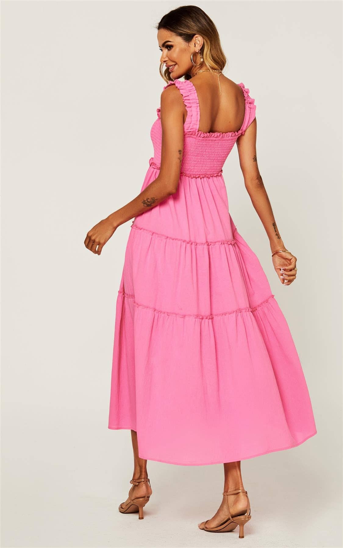 A woman modeling a bright pink tiered midi dress with ruffled shoulder straps and a smocked bodice, shown from the back against a light gray studio background.
