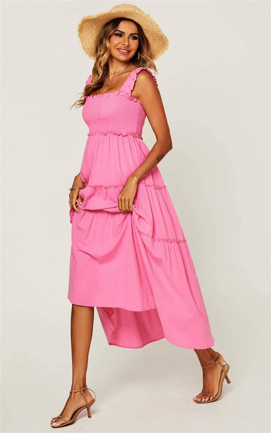 A woman modeling a bright pink tiered midi dress with ruffled shoulder straps, a smocked bodice, and a flared skirt, styled with a straw hat and tan heeled sandals against a light gray studio background.