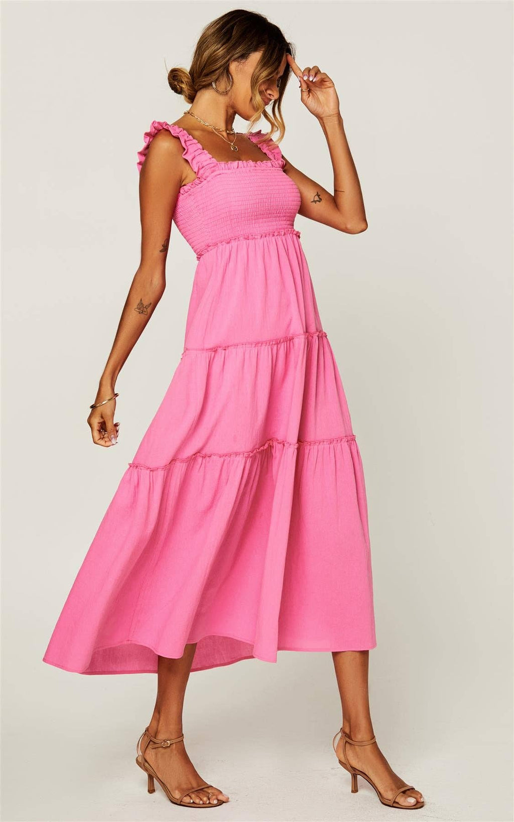 A woman modeling a bright pink tiered midi dress with ruffled shoulder straps, paired with tan heeled sandals, against a light gray studio background.
