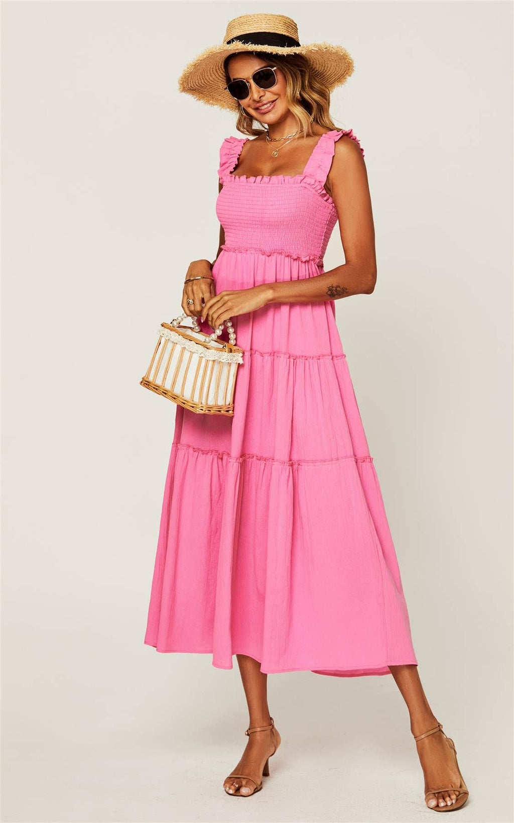 A woman modeling a bright pink tiered midi dress with ruffled straps, styled with a straw hat, sunglasses, a small woven handbag, and tan heeled sandals against a light gray studio background.